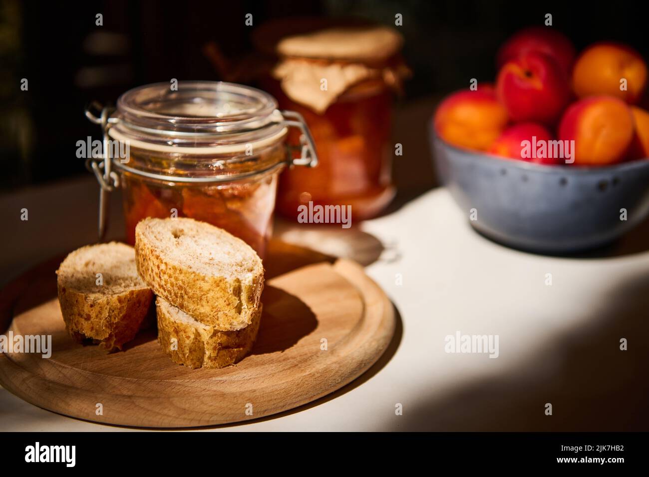 Stillleben. Scheiben Vollkornbrot auf einem Holzbrett, ein Glas mit hausgemachter Marmelade und reifen Aprikosen auf einem weißen Tisch Stockfoto Stillleben. Scheiben Vollkornbrot auf einem Holzbrett, ein Glas mit hausgemachter Marmelade und reifen Aprikosen auf einem weißen Tisch Stockfoto