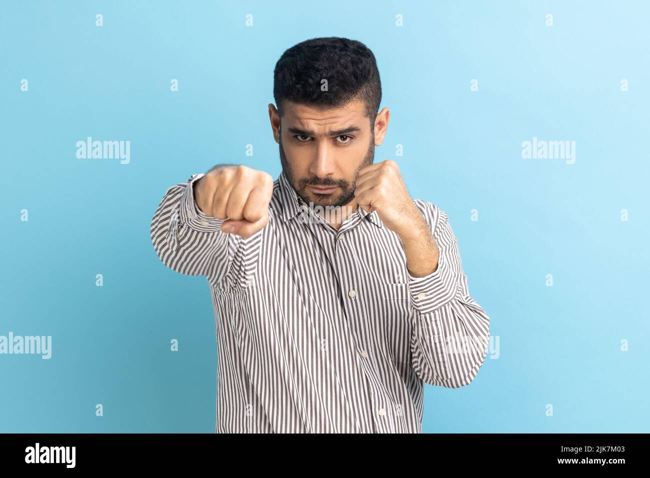 Porträt eines aggressiven jungen erwachsenen Geschäftsmannes, der mit Boxfäusten und bereit zum Angriff oder zur Verteidigung steht, mit einem wütenden Gesicht aussieht und ein gestreiftes Hemd trägt. Innenaufnahme des Studios isoliert auf blauem Hintergrund Stockfoto