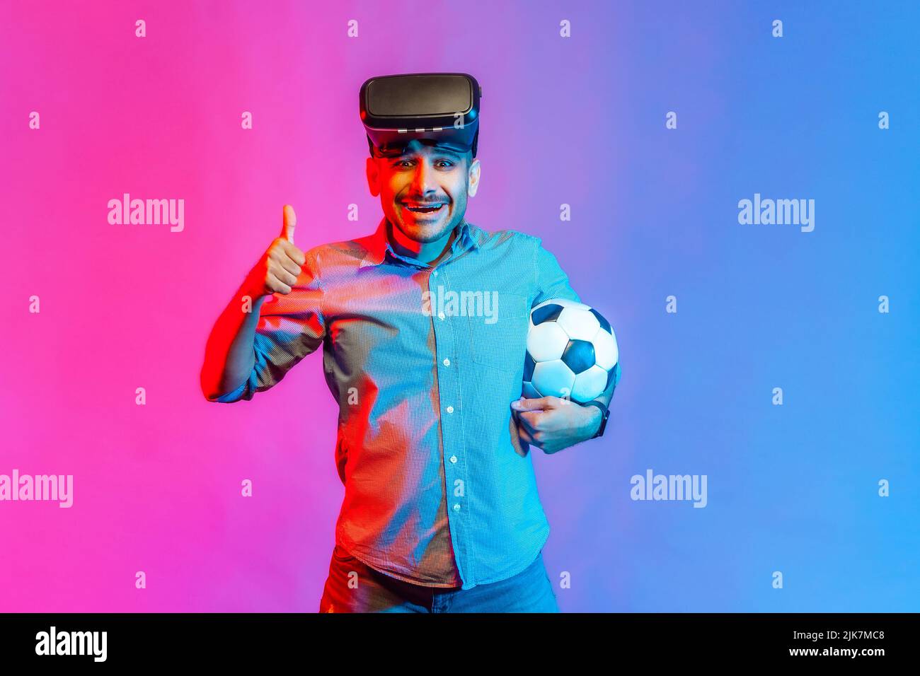 Porträt eines Mannes im Hemd, der eine Virtual-Reality-Brille trägt, mit einem Fußballball steht und den Daumen nach oben zeigt, wie bei einem Fußballspiel. Innenaufnahmen im Studio, isoliert auf farbigem Neonlicht-Hintergrund. Stockfoto