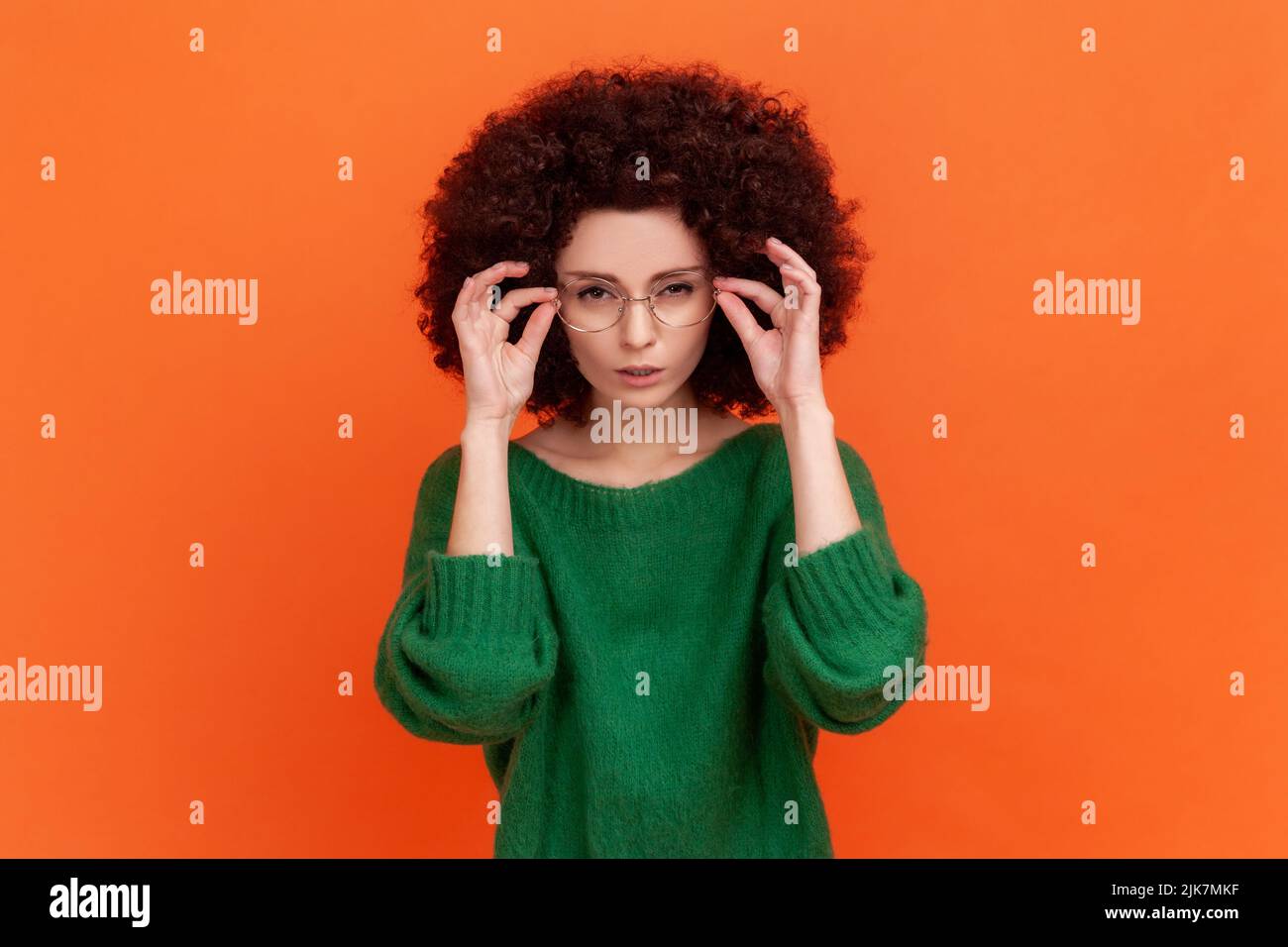 Porträt einer aufmerksamen schönen Frau mit Afro-Frisur tragen grünen lässigen Stil Pullover stehend Brille und Blick auf die Kamera. Innenaufnahme des Studios isoliert auf orangefarbenem Hintergrund. Stockfoto