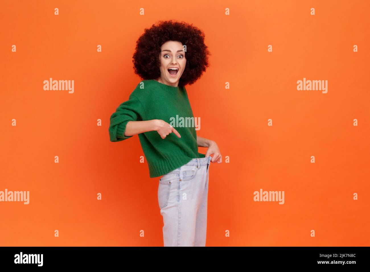 Glückliche Frau mit Afro-Frisur trägt grünen lässigen Stil Pullover zeigt schlanke Taille in großen Hosen, erfolgreiche Gewichtsabnahme, Diät Konzeption. Innenaufnahme des Studios isoliert auf orangefarbenem Hintergrund. Stockfoto