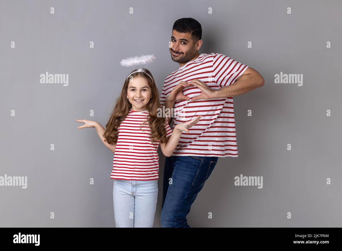 Porträt eines optimistischen Vaters und einer Tochter in gestreiften T-Shirts, die zusammen posieren, eines kleinen Mädchens mit Heiligenschein über dem Kopf, eines Mannes, der mit den Händen eine Herzbewegung zeigt. Innenaufnahme des Studios isoliert auf grauem Hintergrund. Stockfoto