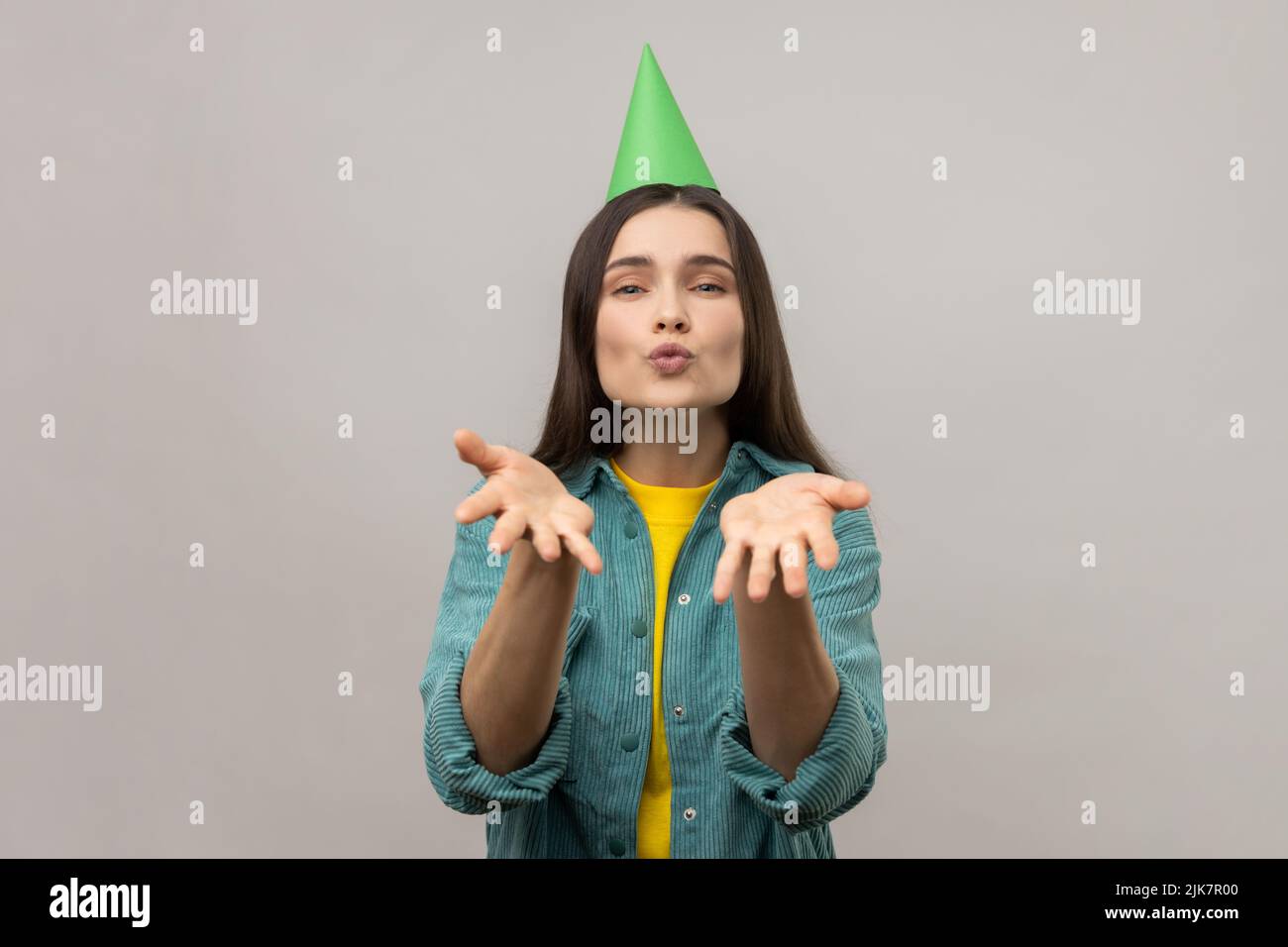 Erfreut Frau mit grünem Kegel senden Luftküsse, feiert ihren Geburtstag, drückt Liebe zu ihren Gästen, tragen lässige Jacke. Innenaufnahme des Studios isoliert auf grauem Hintergrund. Stockfoto