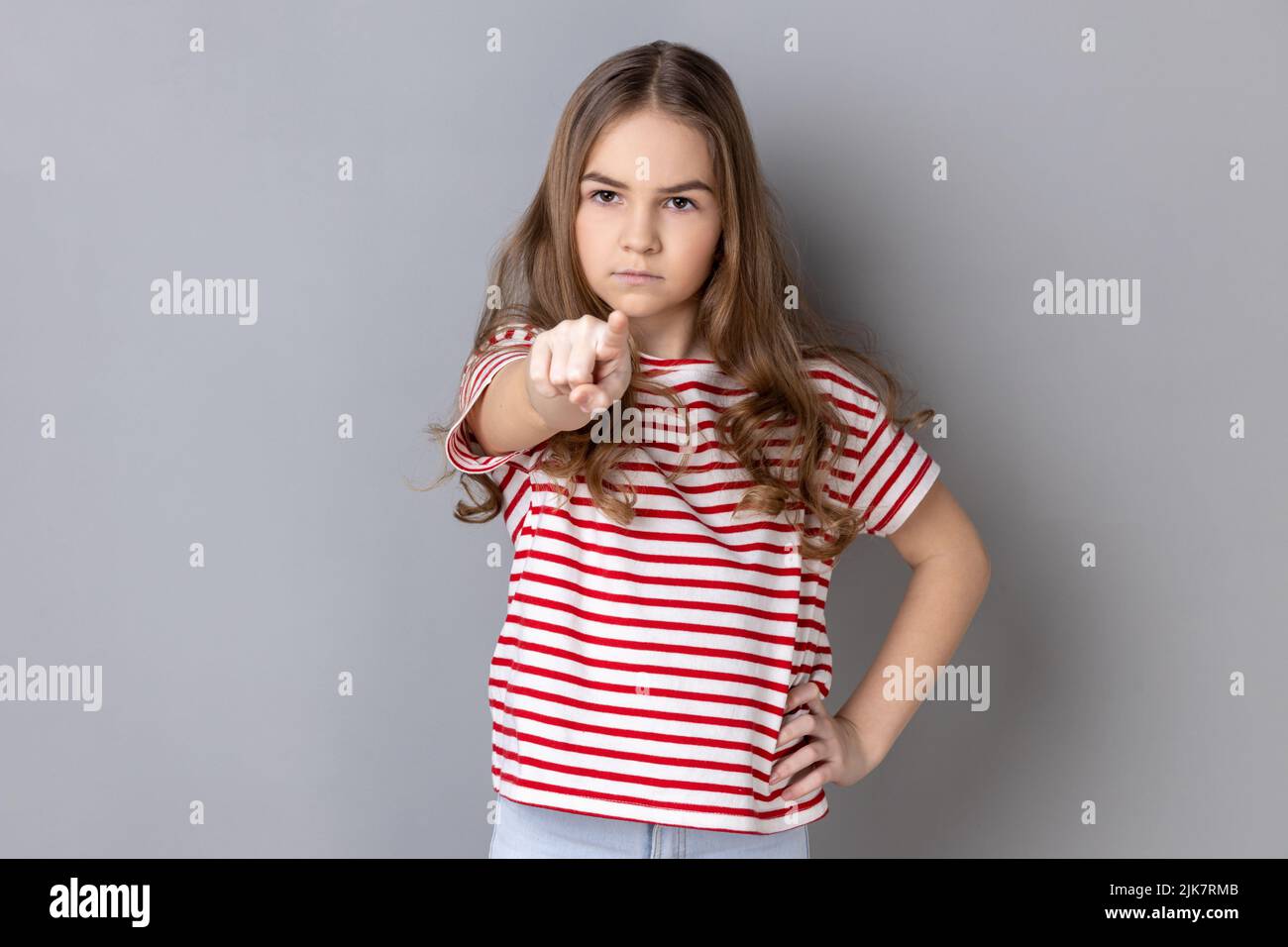Porträt eines streng dunkelhaarigen kleinen Mädchens in gestreiftem T-Shirt, das den Finger auf die Kamera zeigt und sich mit einem schweren, herrischen Gesicht beschuldigt. Innenaufnahme des Studios isoliert auf grauem Hintergrund. Stockfoto