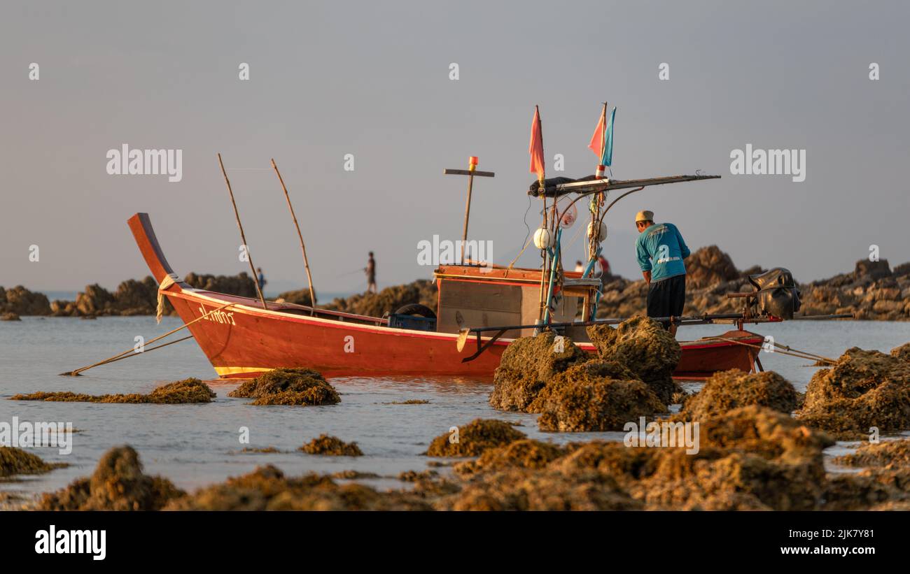 Ein thailändisches Longtail-Boot, das zwischen Felsen am Strand festgemacht wurde und der Kapitän seine Angelausrüstung vorbereitete Stockfoto