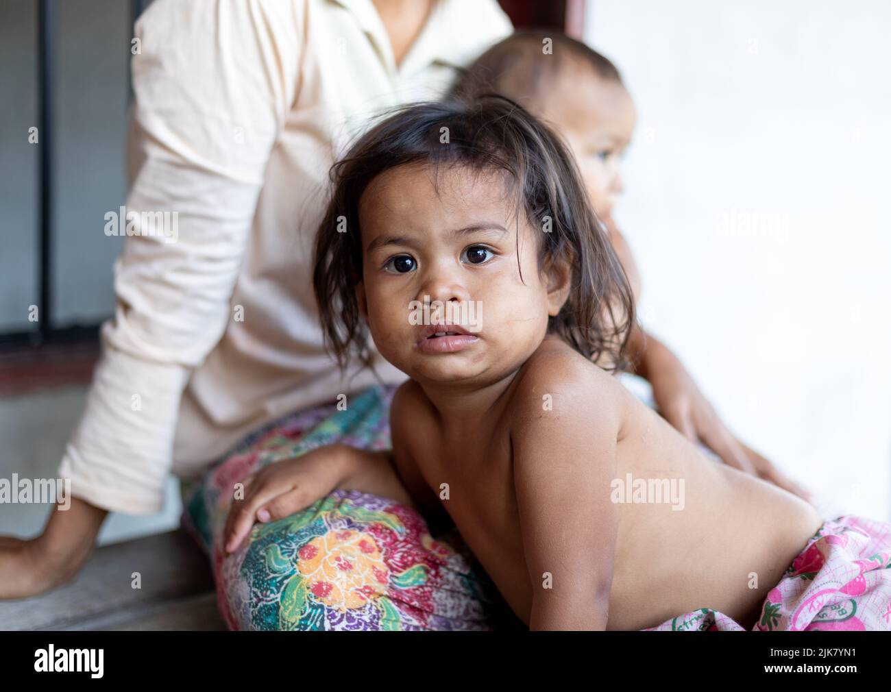 Ein junges Thai-Mädchen mit schmutzigem Gesicht starrt auf die Kamera, während sie sich auf den Schoß ihrer Mutter lehnt Stockfoto