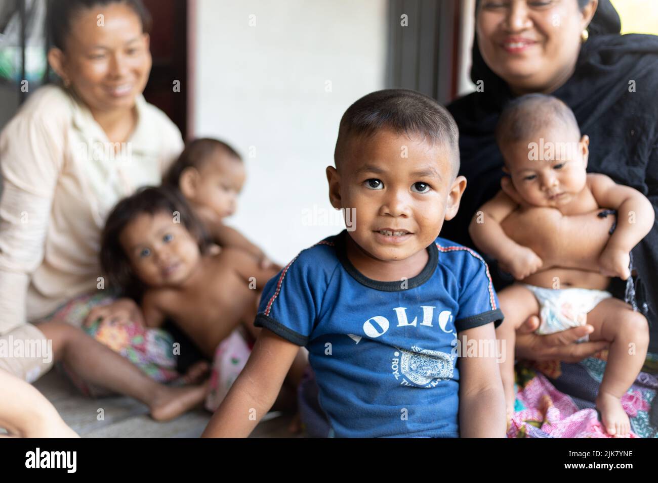 Ein junger Thai-Junge blickt auf die Kamera, und seine lächelnde Familie sitzt dahinter. Bruder, Schwestern, Mutter, Freund. Thailand, arm, ländlich. Stockfoto