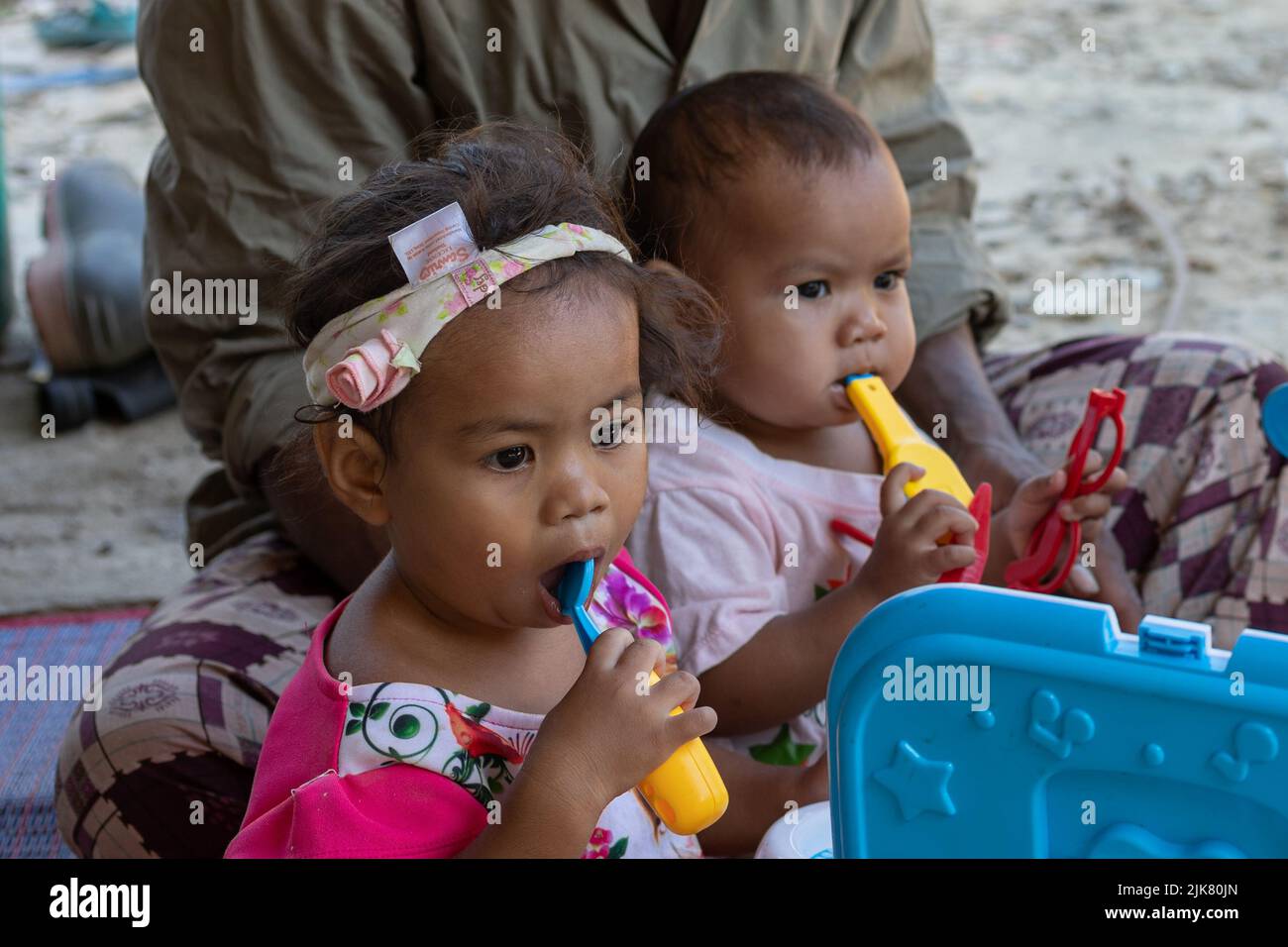 Zwei junge Thai-Mädchen sitzen mit ihrem Vater und halten Plastikspielzeug am Mund Stockfoto