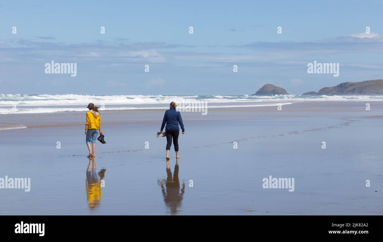 Zwei Damen laufen im nassen Sand an einem sonnigen Tag im Sommer an einem Strand entlang, während die Wellen des Atlantischen Ozeans am Ufer aufprallen. Reflexionen auf nassem Sand Stockfoto
