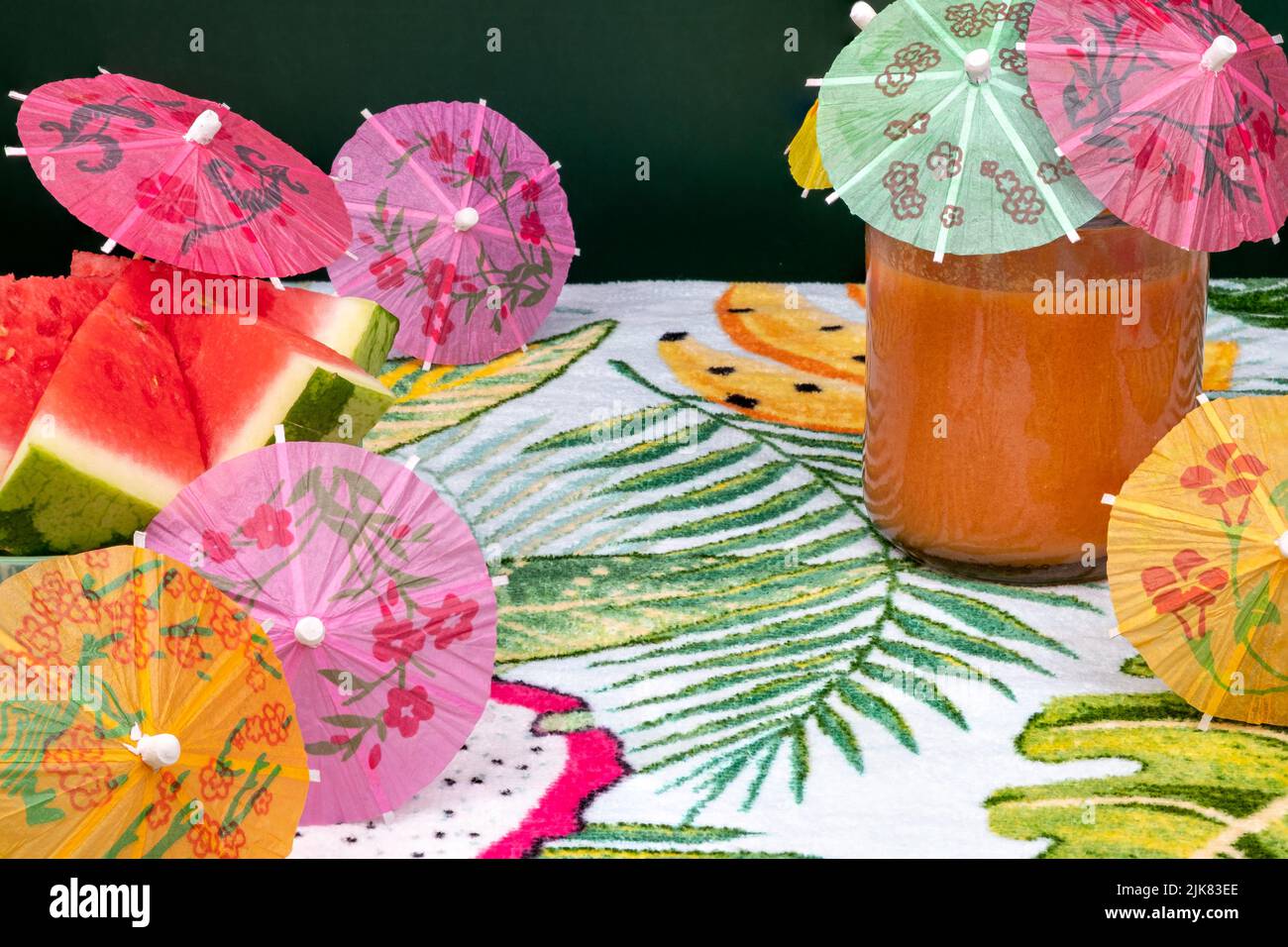 Frisch geschnittene Wassermelone mit einem kleinen Regenschirm, ein Glas Gazpacho auf einem Tisch mit Palmen. Stockfoto