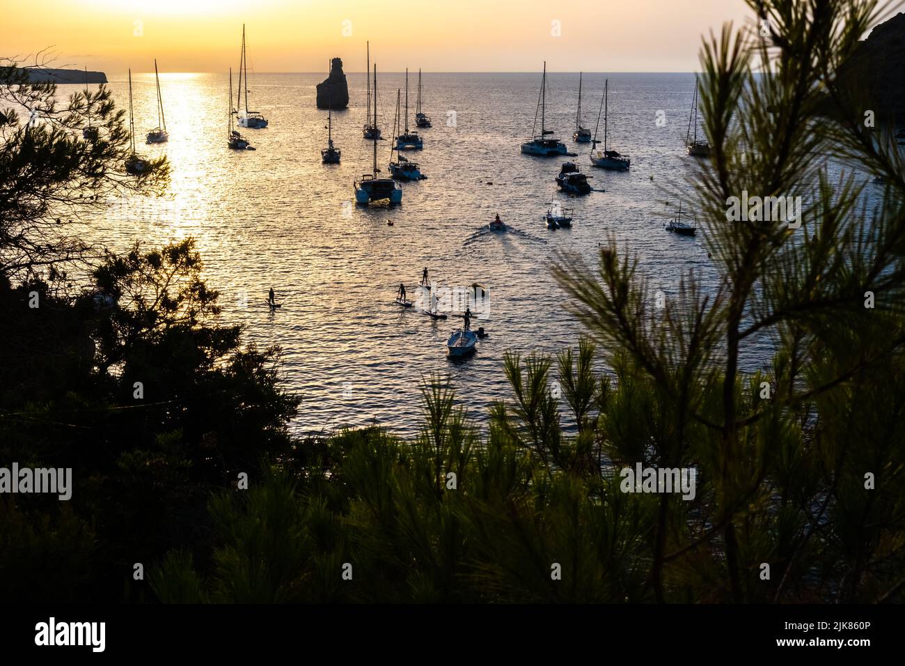 Touristen üben Paddelsurfing zwischen Booten, die in einer Bucht während des Sonnenuntergangs festgemacht sind. Stockfoto