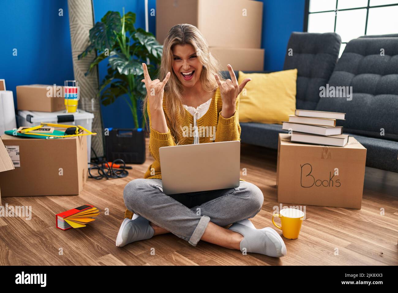 Junge Frau sitzt auf dem Boden in neuen Hause mit Laptop schreiend mit verrücktem Ausdruck Rock-Symbol mit Händen oben. Music Star. Heavy-Konzept. Stockfoto