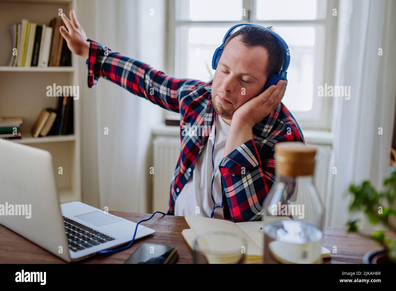 Junger Mann mit Down-Syndrom, der im Büro am Schreibtisch sitzt und mit einem Laptop Musik von Kopfhörern hört. Stockfoto