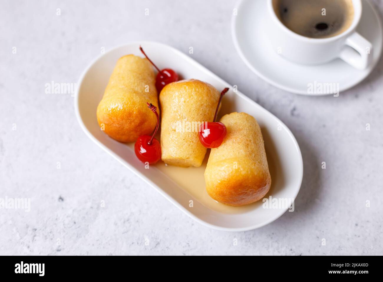 Neapolitanischer Rum baba (oder baba au Rum) auf einem weißen Teller mit einer Cocktailkirsche und einer Tasse Kaffee. Kleine, in Rumsirup getränkte Hefekuchen auf einem grauen Rücken Stockfoto