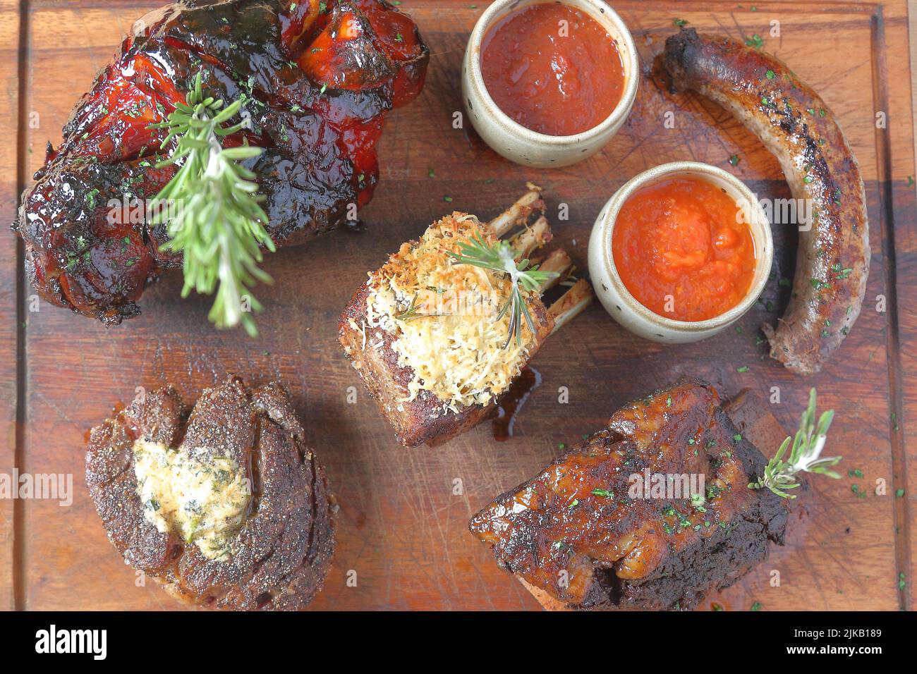 Gemischte Grillplatte mit Braten, Lamm, Schwein und Wurst, garniert mit Saucen auf Tomatenbasis, präsentiert auf einem Holzbrett Stockfoto