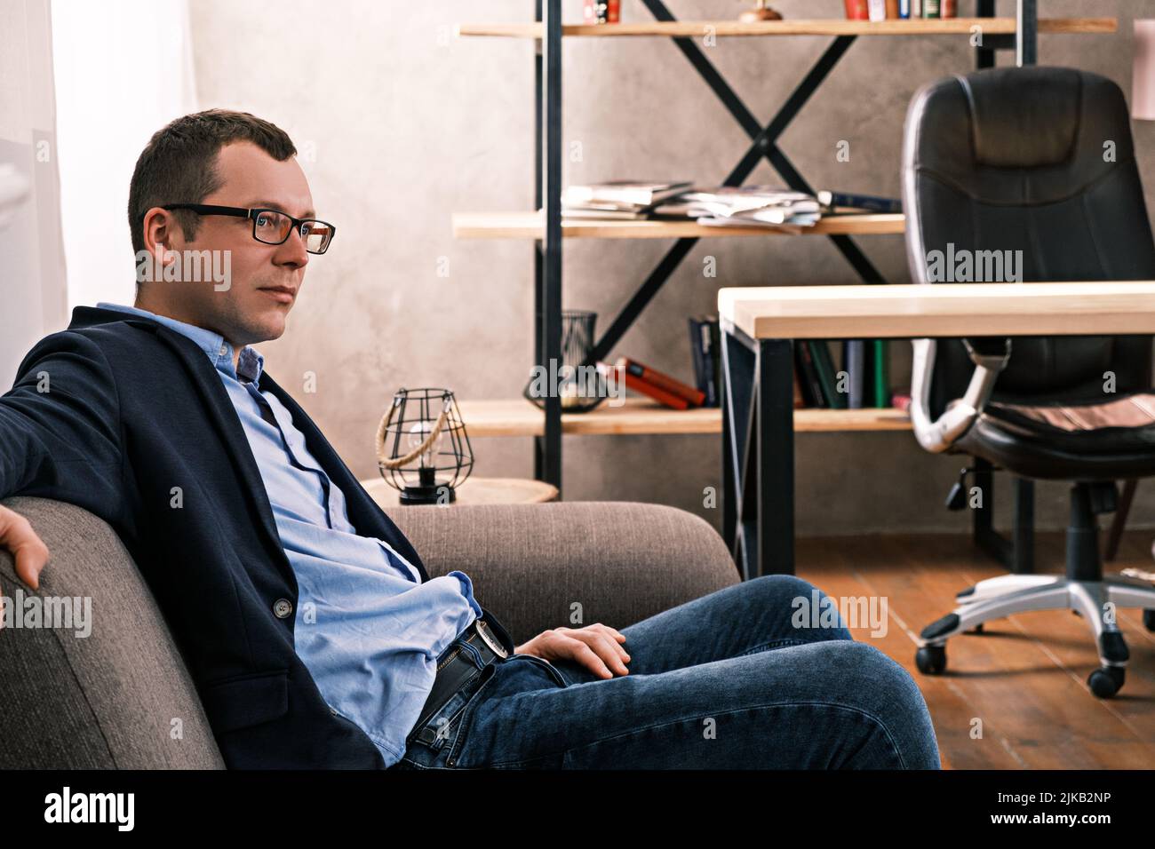 Portrait Seitenansicht des kaukasischen Mannes in Jacke und Brille, der auf dem Sofa sitzt und vor dem Hintergrund eines Fensters und von Regalen mit Dokumenten blickt Stockfoto