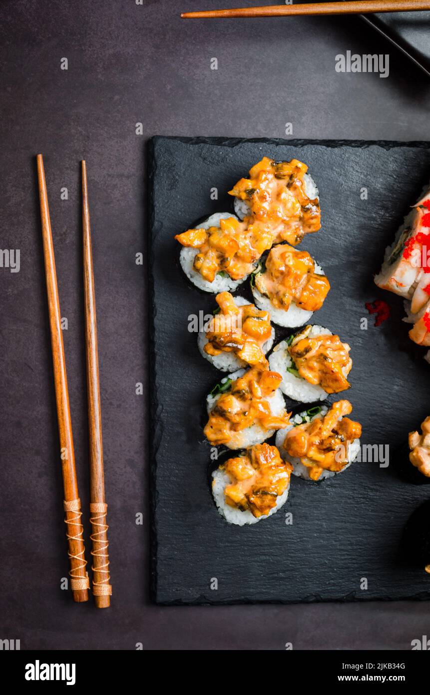 Leckere Brötchen auf Steinplatte mit Saucen, Essstäbchen, Ingwer und Wasabi auf dunklem Tisch. Asiatische Küche. Lieferservice Japanisches Essen . Verschiedene Sushi-Sorten Stockfoto