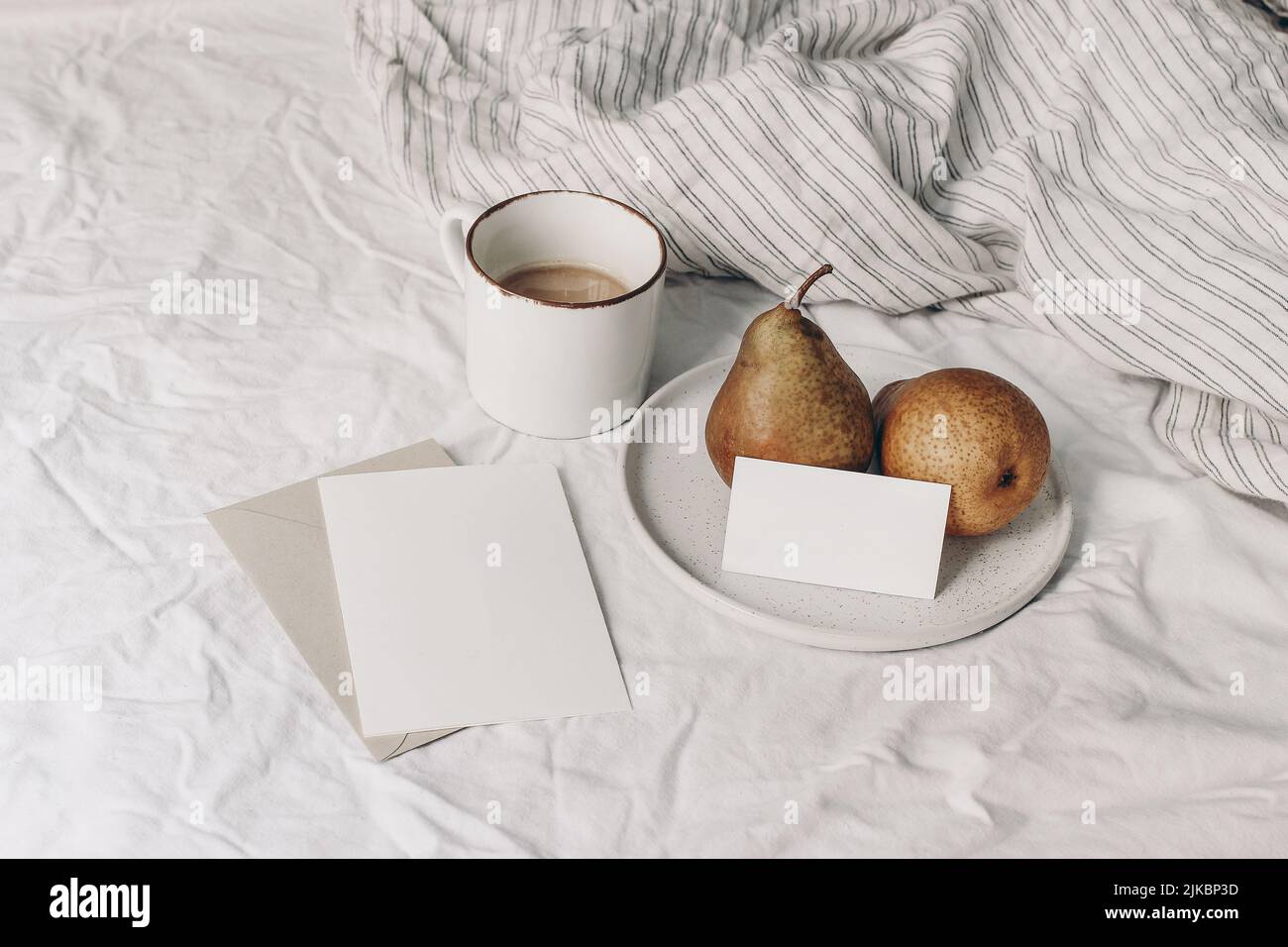 Herbstfrühstück in Bettkomposition. Tasse Kaffee, leere Begrüßung und Visitenkarten-Mockups. Birnenfrüchte auf Keramikplatte. Weiß gestreiftes Leinen Stockfoto