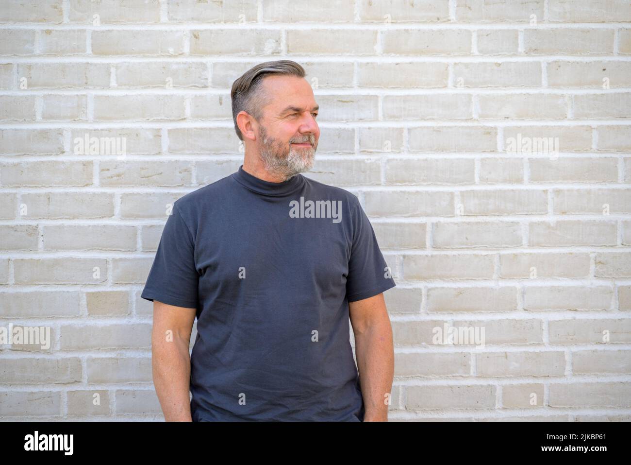 Entspannter bärtiger Mann mittleren Alters im Sommer-T-Shirt, der vor einer Ziegelwand posiert und mit einem Lächeln zur Seite schaut Stockfoto