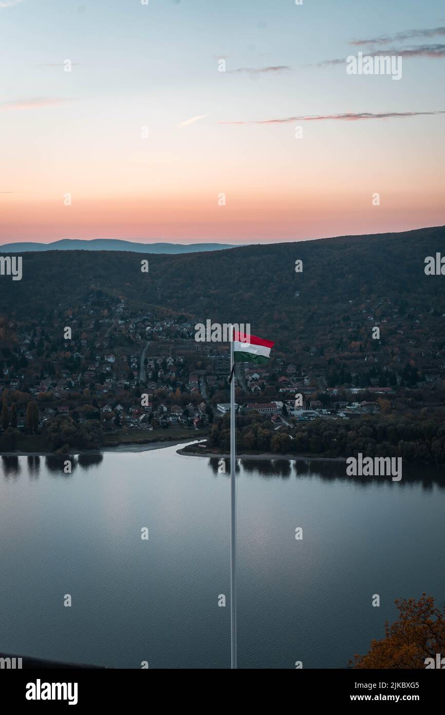 Eine vertikale Aufnahme der ungarischen Flagge auf einem langen Pol an einem See mit Häusern der Stadt und Klippen im Hintergrund Stockfoto