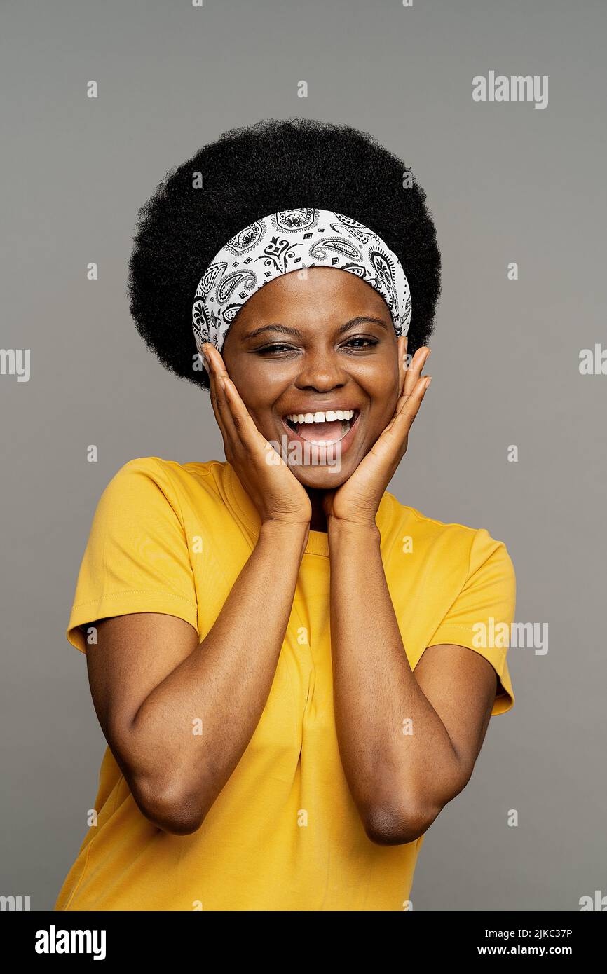 Fröhliche afroamerikanische Frau mit Stirnband Fixierung verkokelte Haare legt die Hände auf das Gesicht lächelnd Stockfoto
