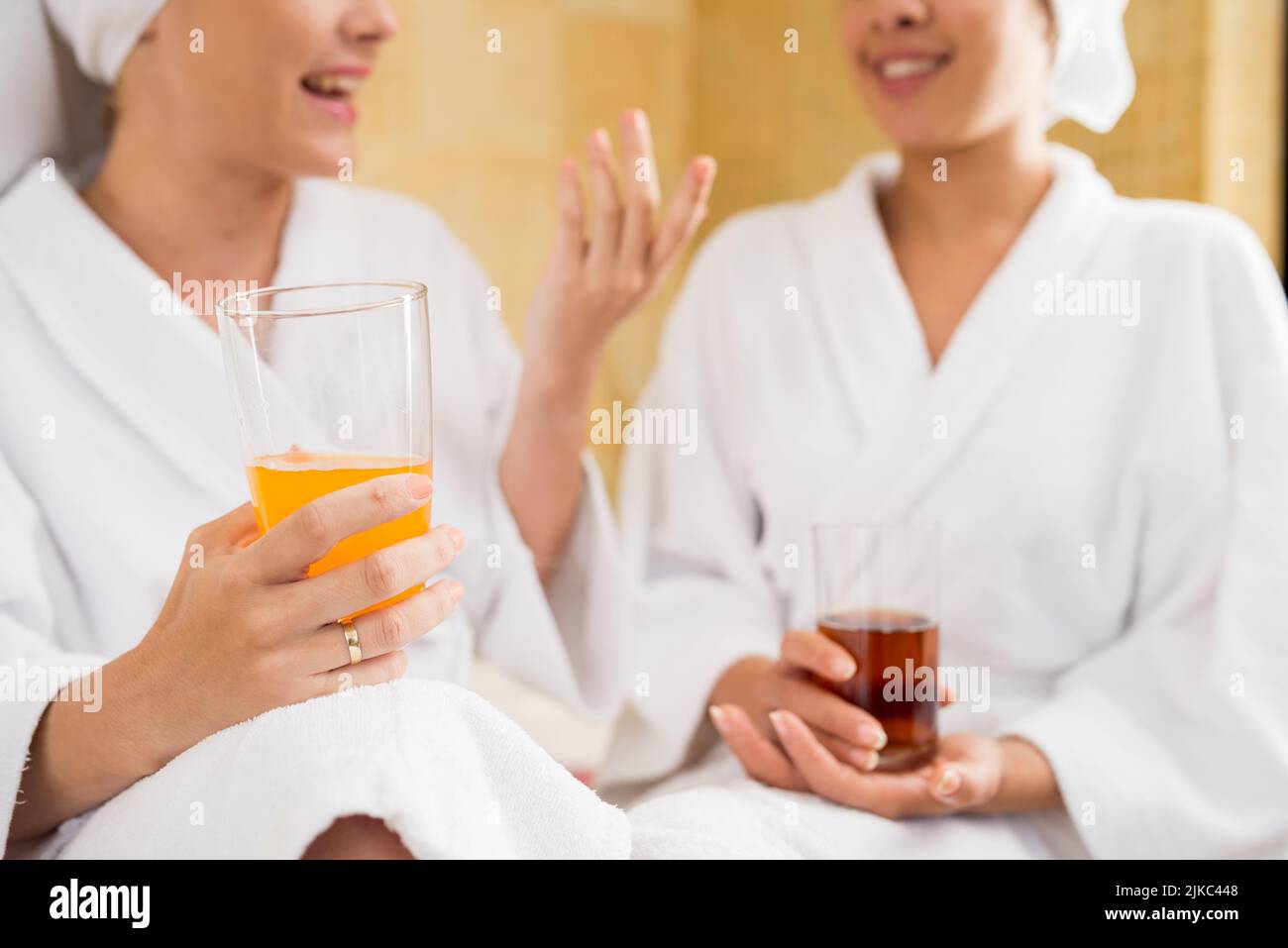 Nahaufnahme von fröhlichen jungen Freunden in Bademänteln, die sich animiert miteinander unterhalten und während des Wochenendes im Wellnesscenter frisch gepressten Saft trinken Stockfoto