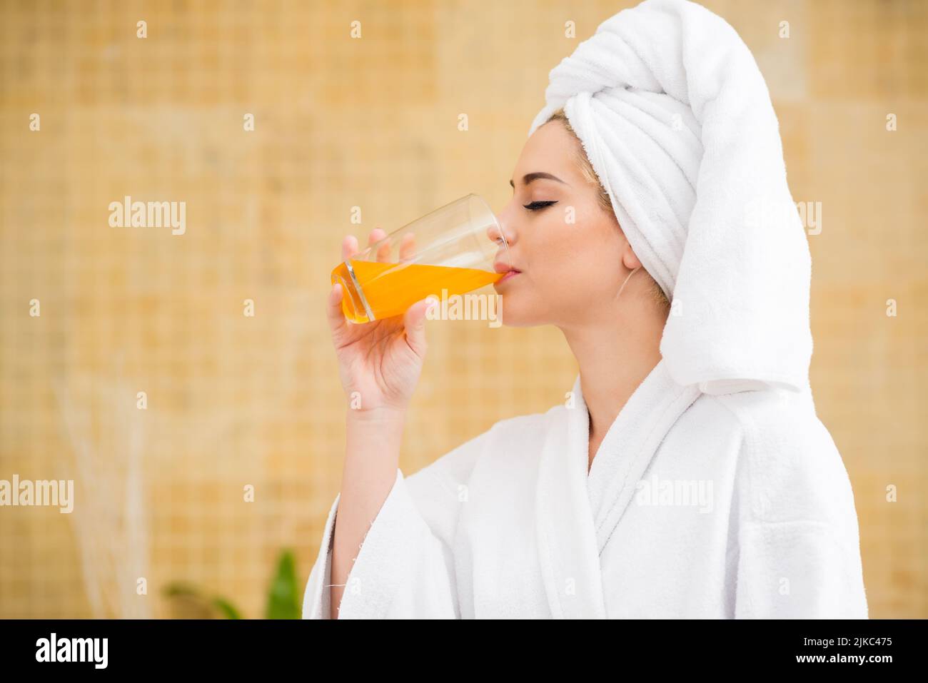Profilansicht einer hübschen jungen Frau, die Bademantel und Handtuch trägt und frisch gepressten Saft trinkt, während sie das Wochenende im Spa-Salon genießt, verschwommener Hintergrund Stockfoto