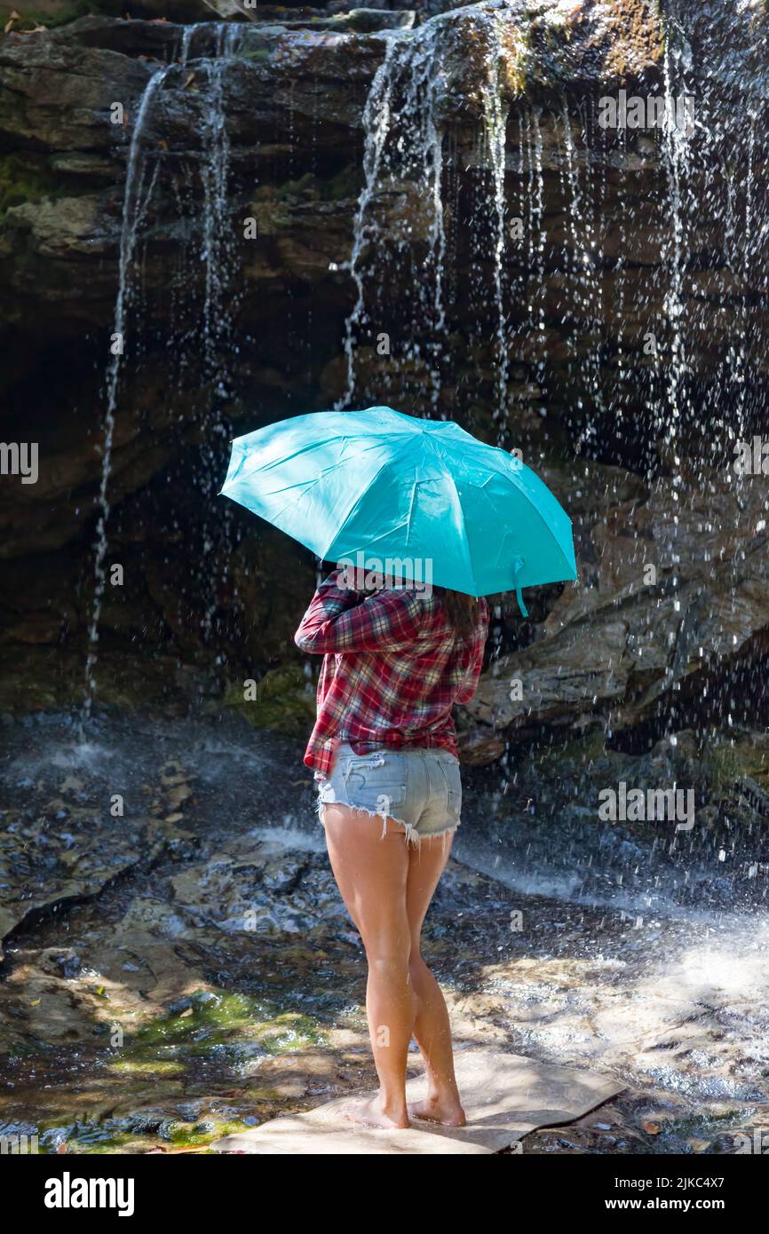 Schöne junge Frau in abgeschnittenen jean-Shorts posiert mit einem türkisfarbenen Regenschirm unter einem Wasserfall auf Big Run Creek in Tucker County, WV. Stockfoto