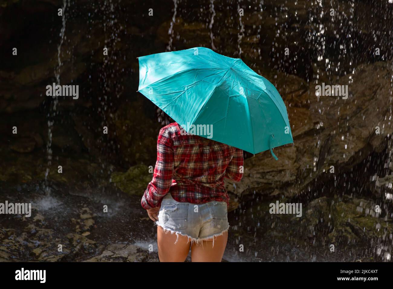 Schöne junge Frau in abgeschnittenen jean-Shorts posiert mit einem türkisfarbenen Regenschirm unter einem Wasserfall auf Big Run Creek in Tucker County, WV. Stockfoto