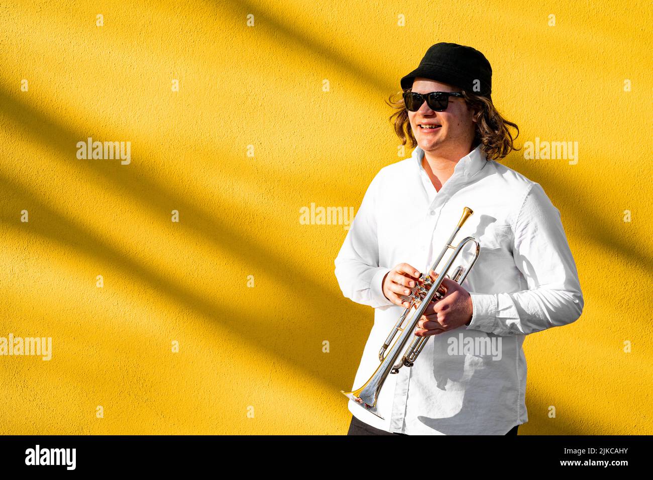 Junger europäischer Musiker in Hemd, Hut und Sonnenbrille, der Trompete spielt Stockfoto