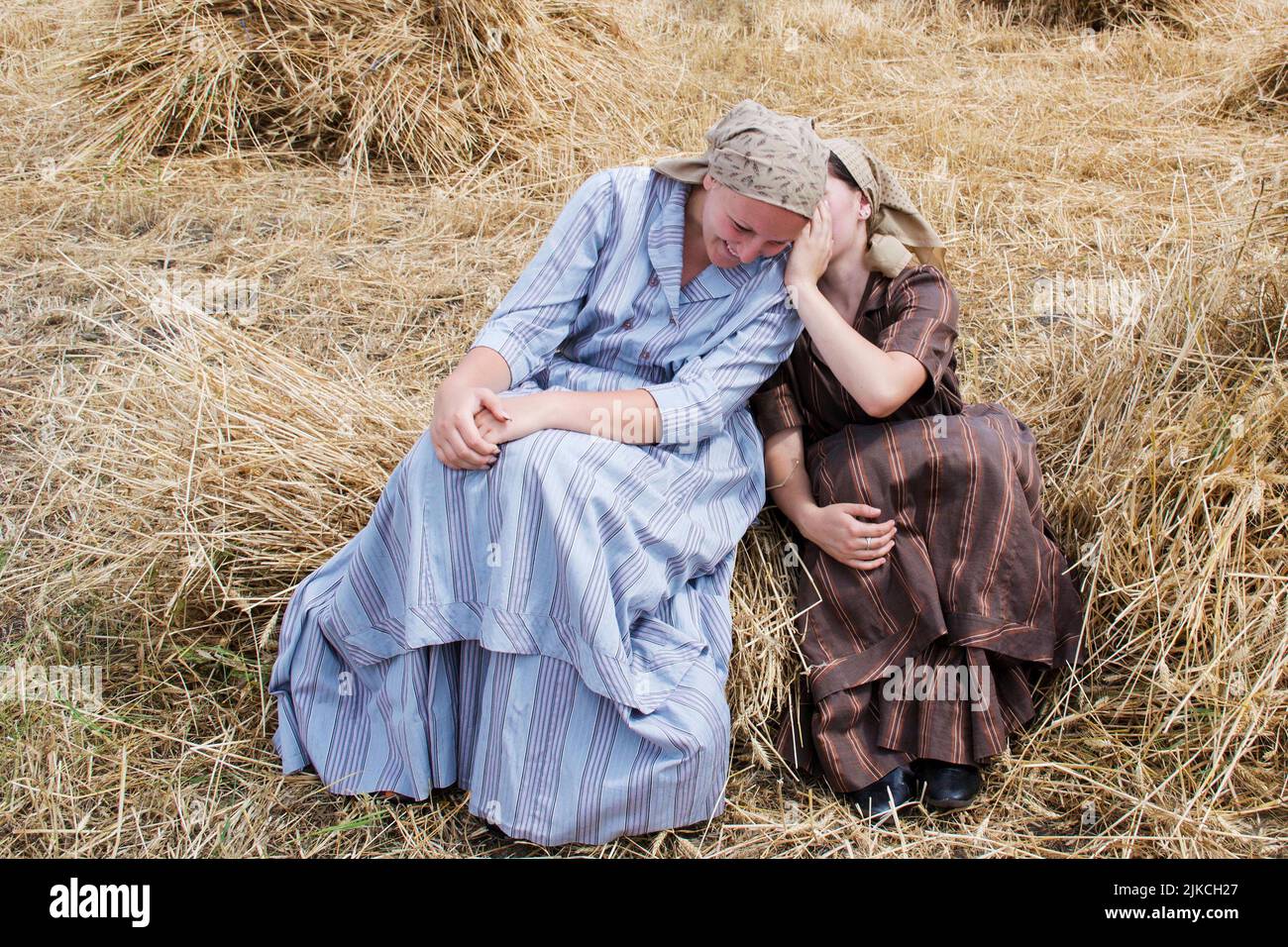 Zwei Frauen in traditionellen serbischen Kleidern sitzen auf einem Stapel getrockneten Heus und flüstern sich gegenseitig zu Stockfoto