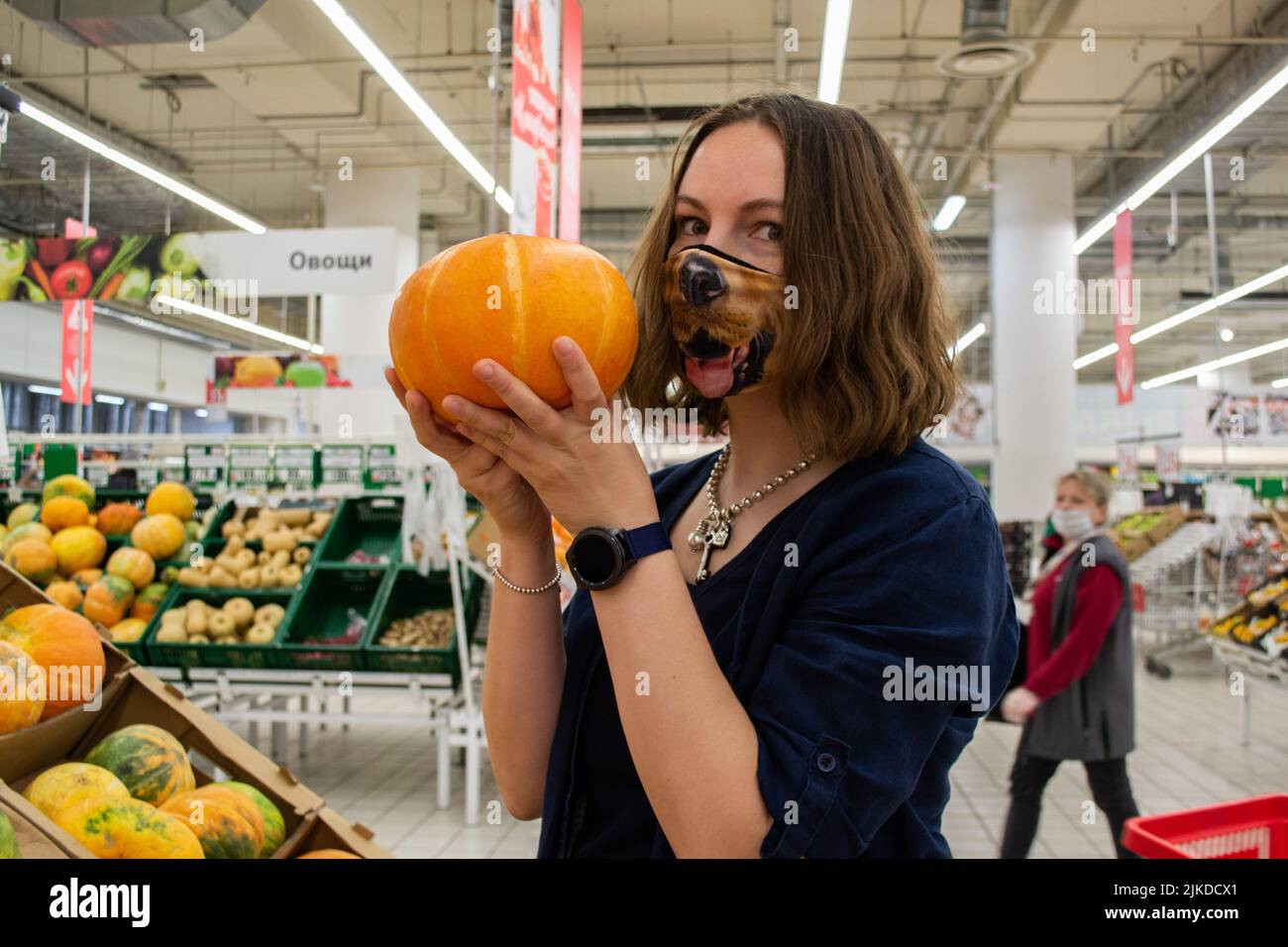 Eine junge Frau in einer lustigen Maske mit einem Hundegesicht hält einen leuchtend orangefarbenen Kürbis in einem Lebensmittelgeschäft. Das Konzept des Schutzes vor dem Virus und bereit für Stockfoto