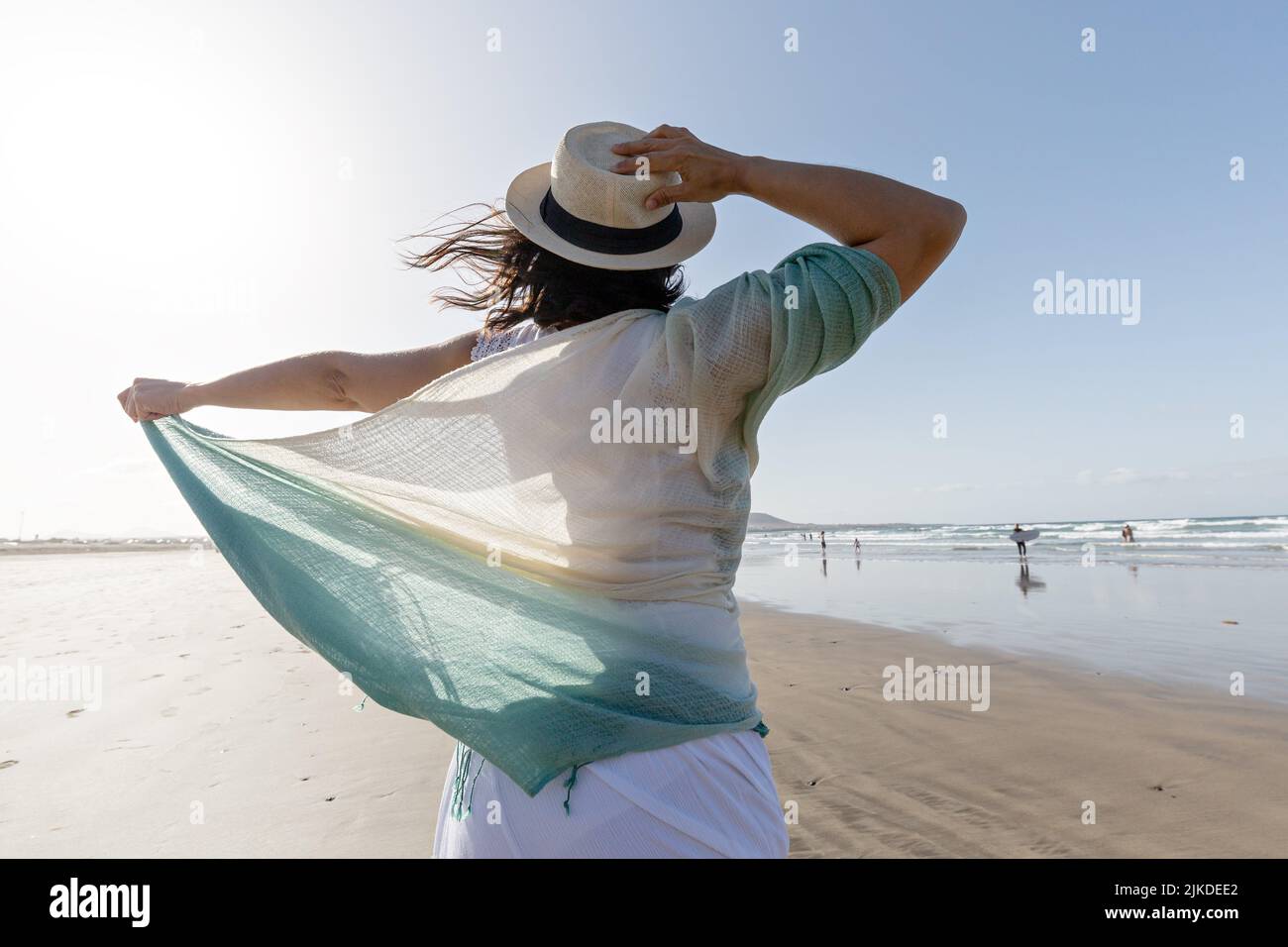 Rückansicht der Brünette mit dünnem Schal berühren Strohhut und genießen die Freiheit, während windigen Wochenendtag auf Sandstrand in der Nähe des Meeres zu verbringen. Stockfoto