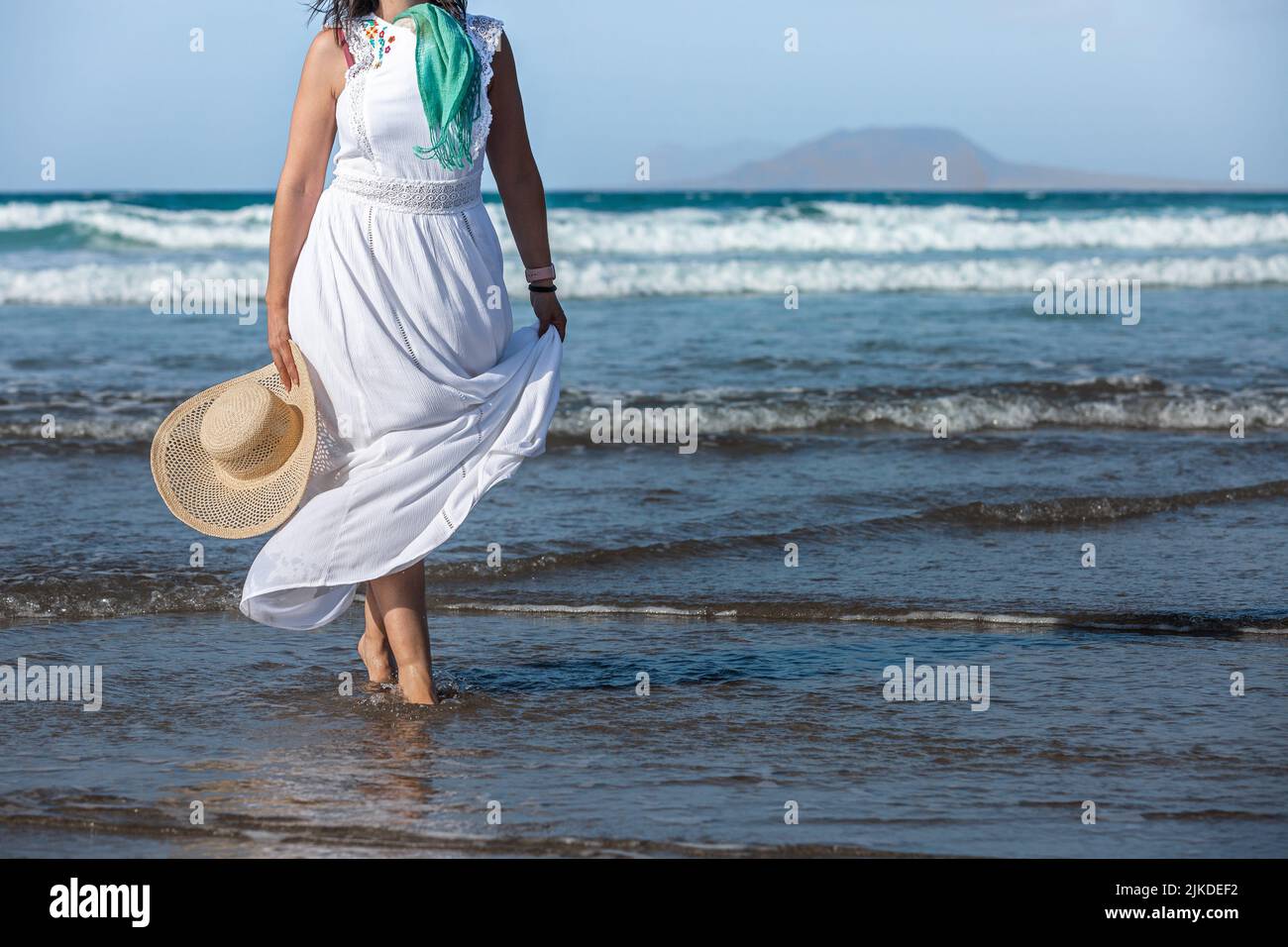 Anonyme Frau in weißem, langem Kleid mit Seidenschal und Strohhut, die während des Sommerurlaubs im Resort in den Wellen schlendert. Stockfoto