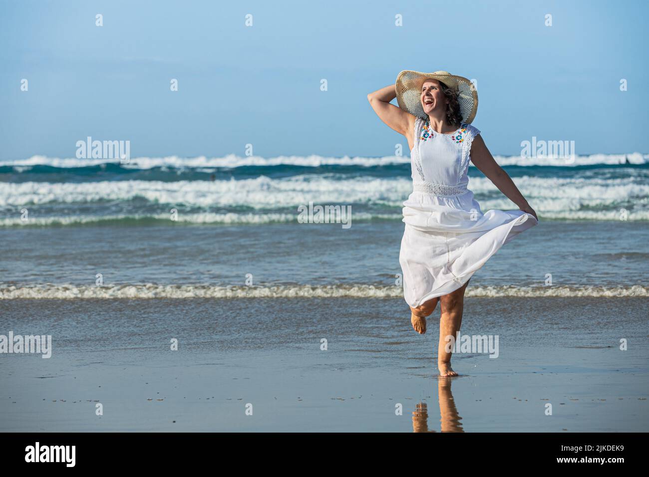 Optimistische weibliche Reisende in weißem Kleid, lachend und wegblickend, während sie auf nassem Sand gegen das winkende Meer und den blauen Himmel rannte. Stockfoto