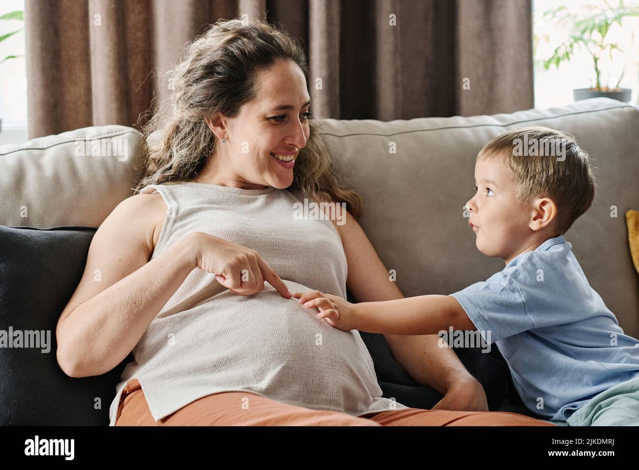 Der entzückende kleine Junge und seine Schwangere zeigen auf ihren Bauch, während sie zusammen spielen und über neues menschliches Leben im Inneren diskutieren Stockfoto