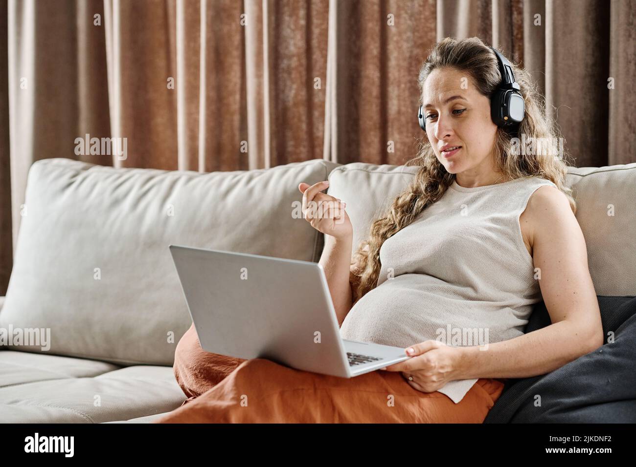 Junge, selbstbewusste, Schwangere mit Kopfhörern, die zu Hause im Video-Chat mit jemandem kommunizieren und dabei auf den Laptop-Bildschirm blicken Stockfoto