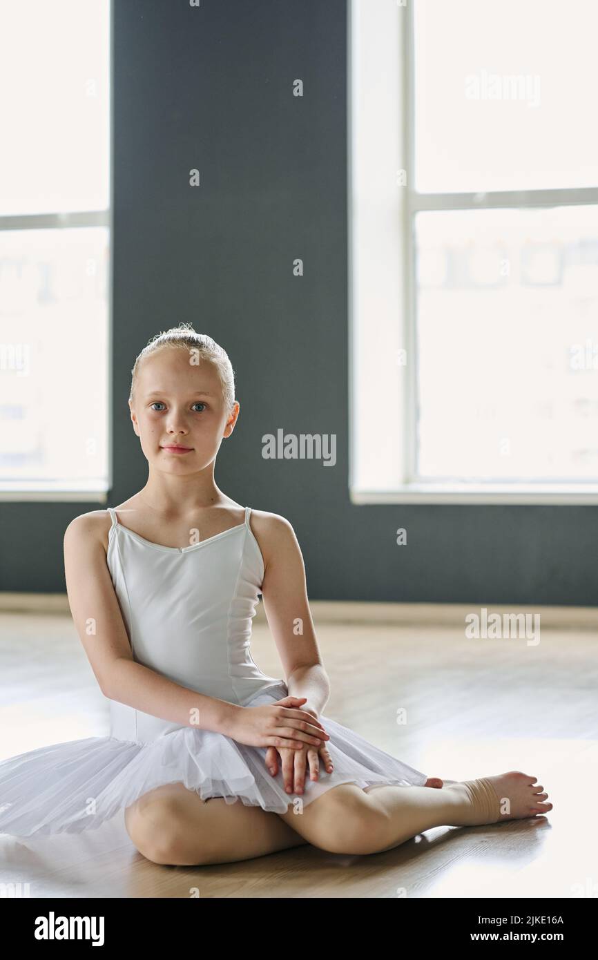 Ziemlich jugendliches Mädchen in weißem Tutu und beigen Strumpfhosen, die auf dem Boden eines geräumigen Klassenzimmers oder Tanzstudios sitzen und die Kamera betrachten Stockfoto