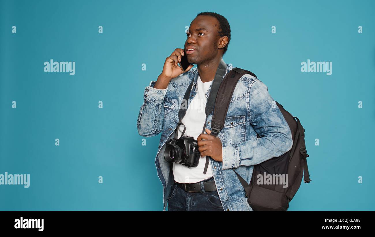 afroamerikanischer Fotograf im Gespräch mit dem Smartphone, Vorbereitung auf Urlaub Reise mit Rucksack und nehmen professionelle Bilder. Ferngesteuerte Unterhaltung vor der Aufnahme von urbane Abenteuerfotos. Stockfoto