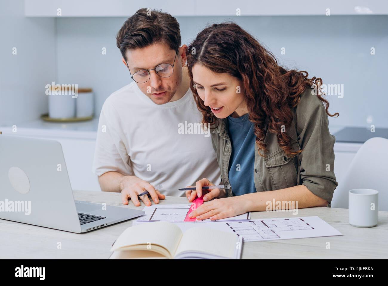 Das Paar sitzt in der Küche, trinkt Kaffee zum Frühstück und spricht über Zeichnungen von Wohnungsrenovierungen, die Notizen in Laptop und Notebook machen Stockfoto