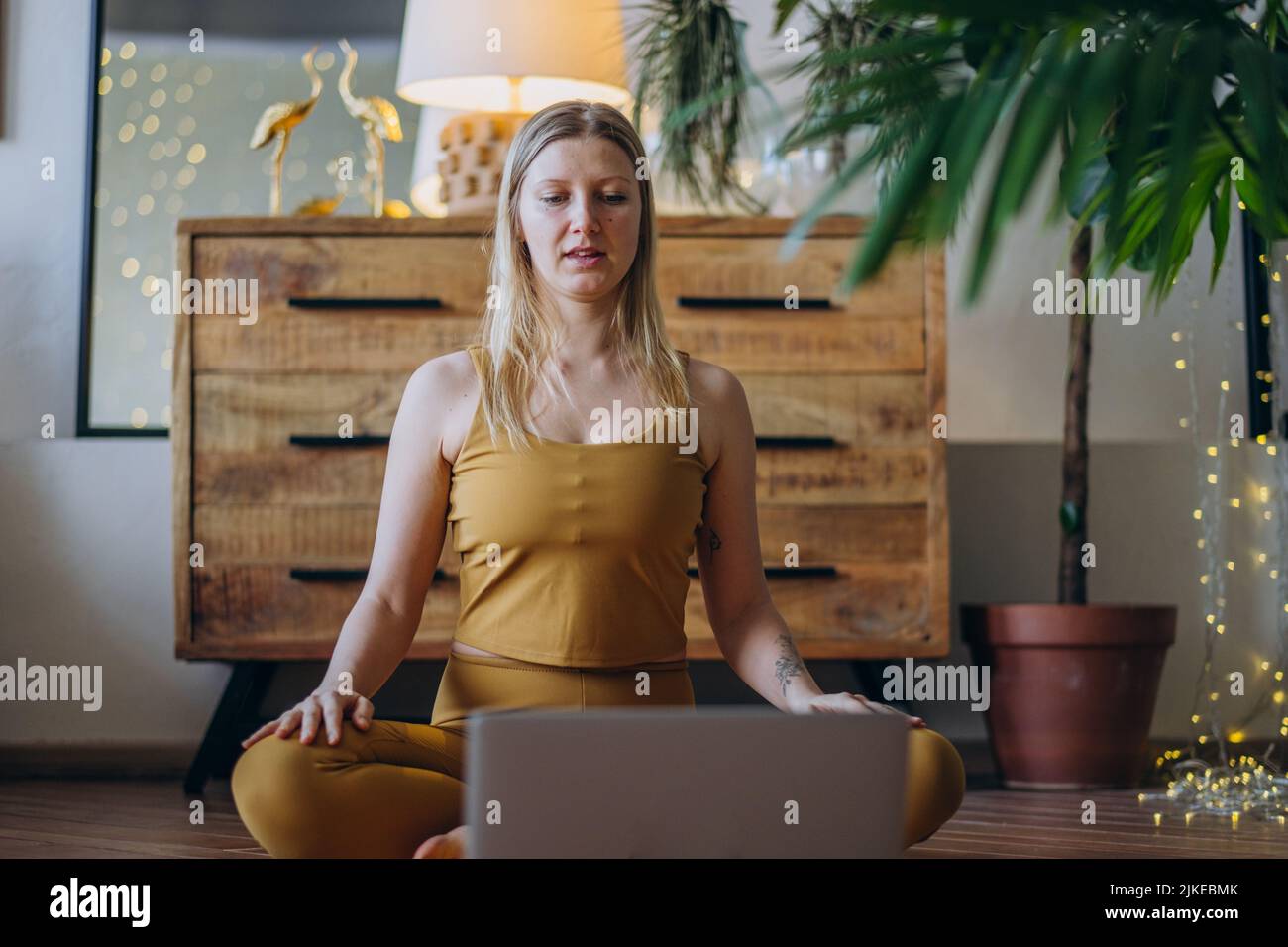Junge Frau in der Oberseite und Leggings macht Dehnübungen auf Yoga-Matte beobachten Tutorial auf Laptop gegen aufhellende Girlande Stockfoto