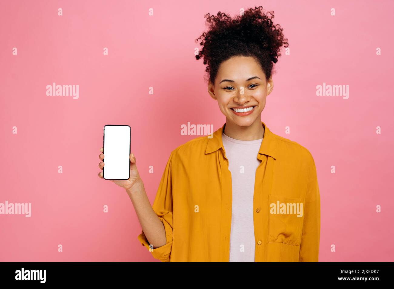 Positive schöne afroamerikanische junge Frau mit lockigem Haar, Mobiltelefon mit leerem weißen Bildschirm in der Hand haltend, Kopierraum für Werbung, freundlich lächelnd, stehend über einem isolierten rosa Hintergrund Stockfoto