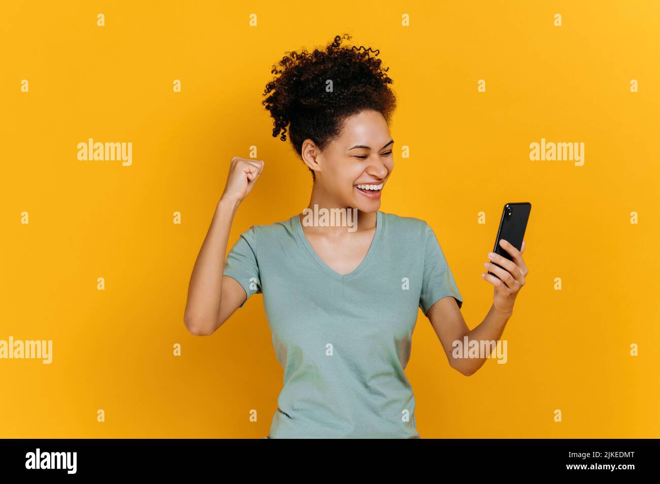 Erstaunt fröhlich aufgeregt afroamerikanischen Mädchen hält Smartphone, unerwartete Nachrichten, Lotterie gewinnen, steht auf isolierten orangen Hintergrund, fröhlichen Gesichtsausdruck, toothy Lächeln, gestikulieren mit Faust Stockfoto