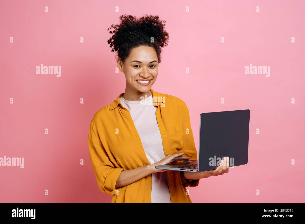 Eine ziemlich erfolgreiche, positive afroamerikanische junge, lockige Frau, die einen offenen Laptop in der Hand hält, sieht freundlich auf die Kamera aus, während sie vor einem isolierten rosa Hintergrund steht und glücklich lächelt Stockfoto