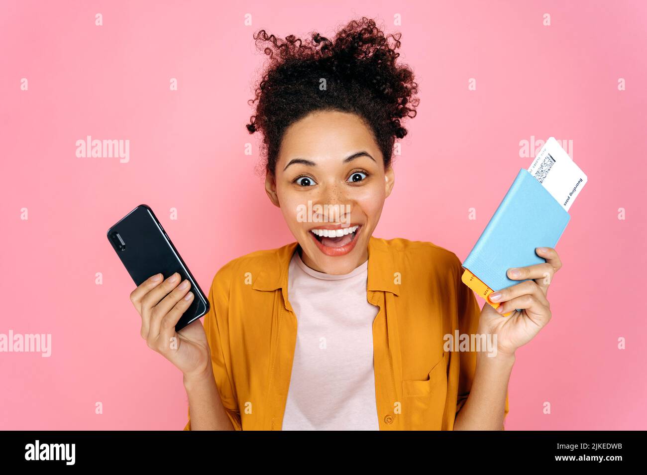 Erstaunt aufgeregt afroamerikanische junge Frau, weibliche Reisende Tourist, in Freizeitkleidung, hält Smartphone und Pass mit Tickets, stehen auf isolierten rosa Hintergrund, schockiert Blicke auf die Kamera, lächelnd Stockfoto