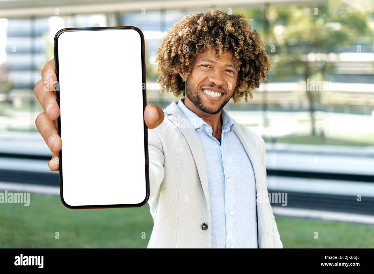 Positiv attraktiver afroamerikanischer oder brasilianischer lockiger Mann, steht im Freien, zeigt Smartphone mit leerem weißen Mockup-Bildschirm mit Platz für Werbung, Präsentation, schaut auf Kamera, lächelt Stockfoto