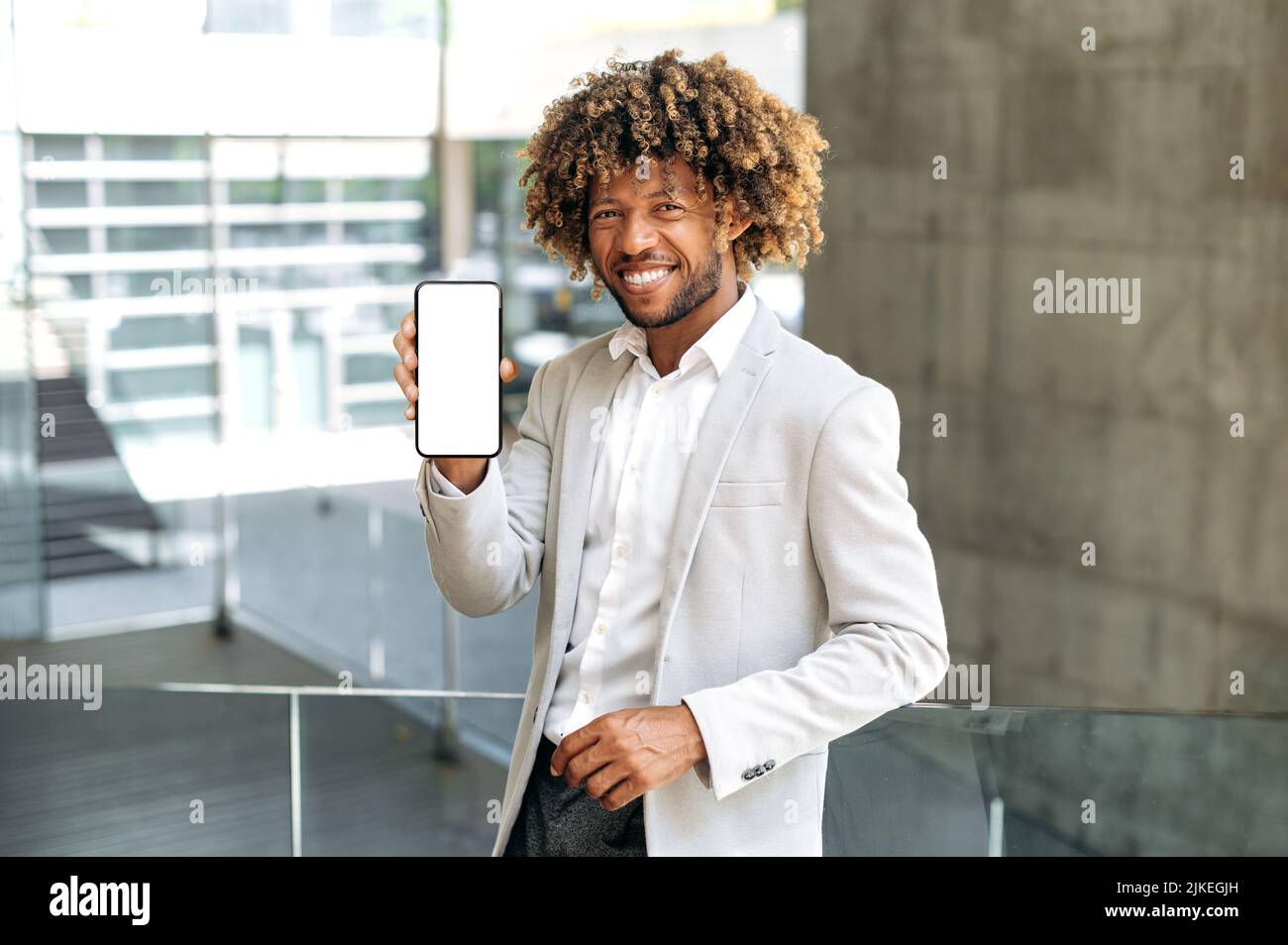 Erfolgreicher, freundlicher afroamerikanischer oder brasilianischer lockiger Mann, steht im Freien in stilvoller Kleidung, zeigt Smartphone mit weißem Mockup-Bildschirm, Platz für Werbung, schaut auf die Kamera, lächelt Stockfoto