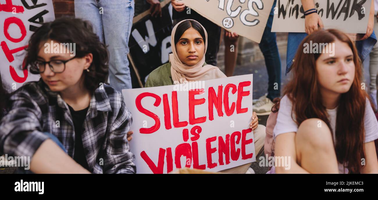 Das muslimische Mädchen blickt auf die Kamera, während es ein Anti-Gewalt-Banner hält. Multiethnische Jugendliche protestieren gegen Krieg und ethnische Gewalt. Jugend aktiv Stockfoto