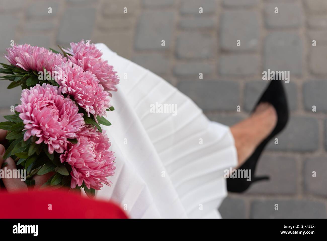 Bouquet von dunkelrosa Astern in den Händen einer sitzenden Frau in einem weißen Faltenrock und schwarzen Stilettos, Draufsicht. Konzept der Entspannung und Reflexion. Stockfoto