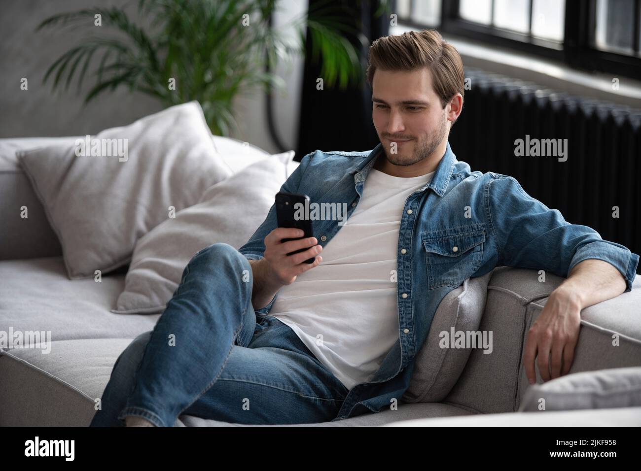 Attraktiver, lächelnder junger Mann in legerer Kleidung, der auf einer Couch im Wohnzimmer sitzt und ein Mobiltelefon benutzt Stockfoto